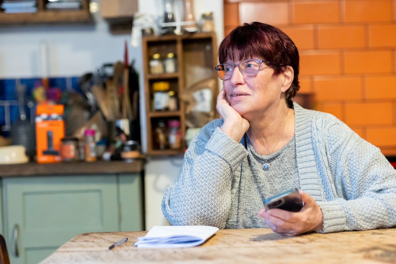A woman at her kitchen table with her phone and a stack of papers